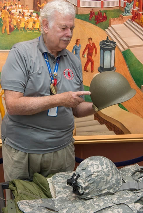 Docent with Military Innovations Discover Cart, holding helmet and showing assorted military body armor.